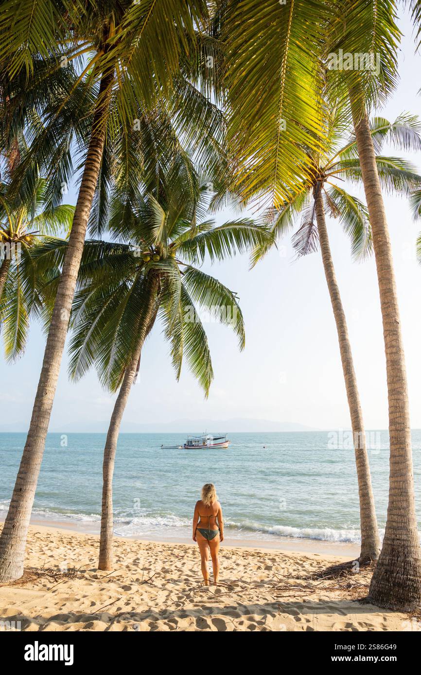 Jeune femme blonde debout sous les palmiers et profitant du soleil du matin à la plage tropicale de Maenam sur Koh Samui, Thaïlande. Banque D'Images