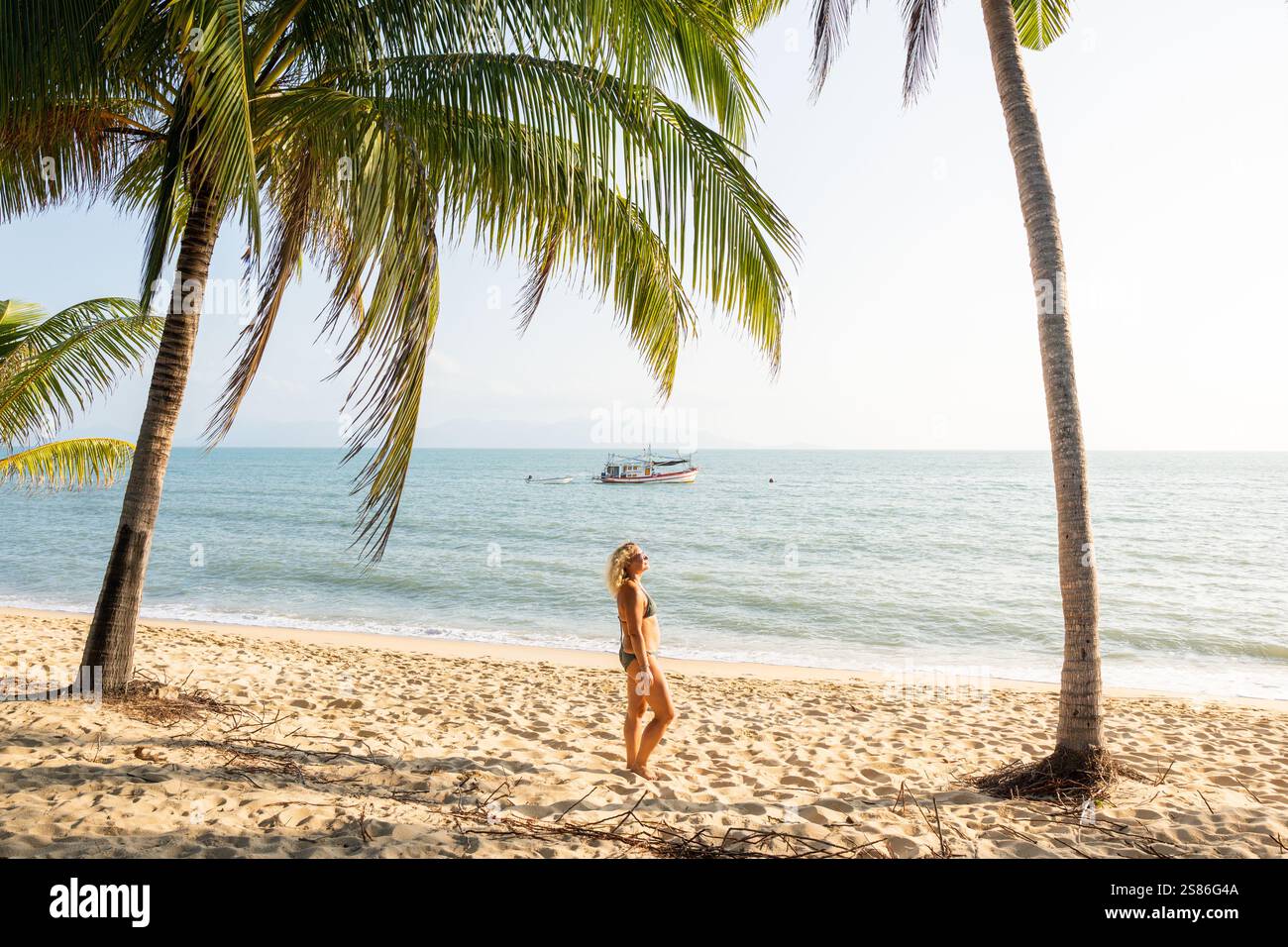 Jeune femme blonde debout sous les palmiers et profitant du soleil du matin à la plage tropicale de Maenam sur Koh Samui, Thaïlande. Banque D'Images