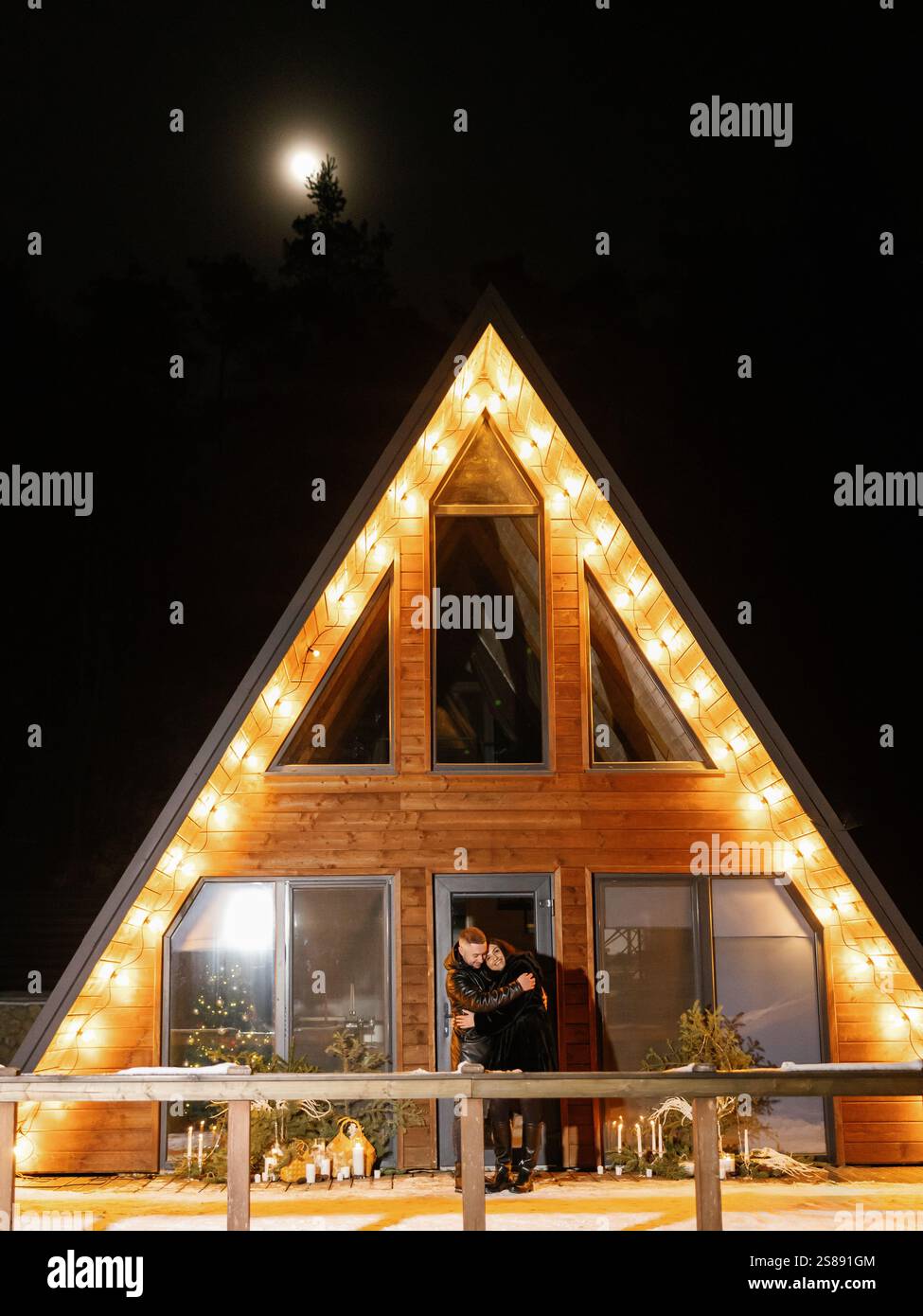 Couple romantique embrassant devant leur chalet décoré de lumières pendant les vacances d'hiver Banque D'Images