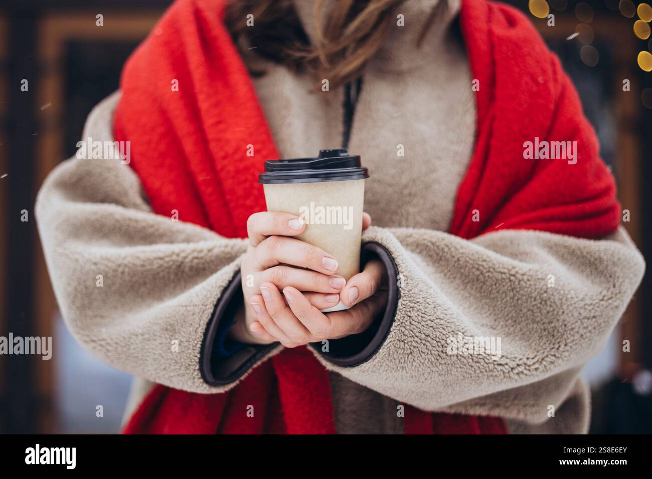 Jeune femme recadrée méconnaissable vêtue d'un manteau en peluche confortable et d'une écharpe rouge frappante tient une tasse à café à emporter, gardant au chaud pendant une journée d'hiver Banque D'Images