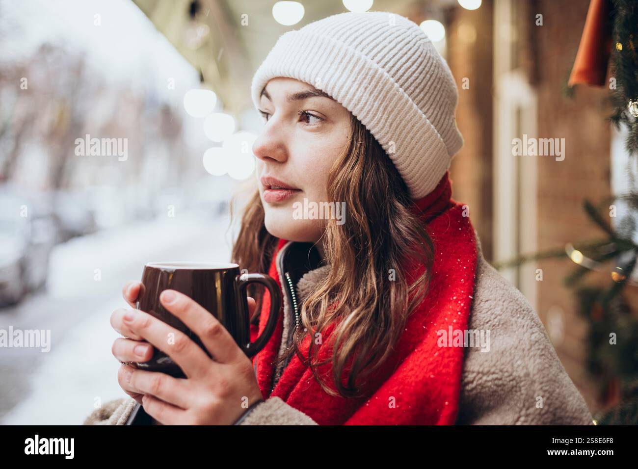 Une jeune femme sirote un chocolat chaud un jour d'hiver enneigé. Vêtue chaudement d'un bonnet blanc et d'une écharpe rouge, elle regarde soigneusement tout en tenant sa tasse Banque D'Images