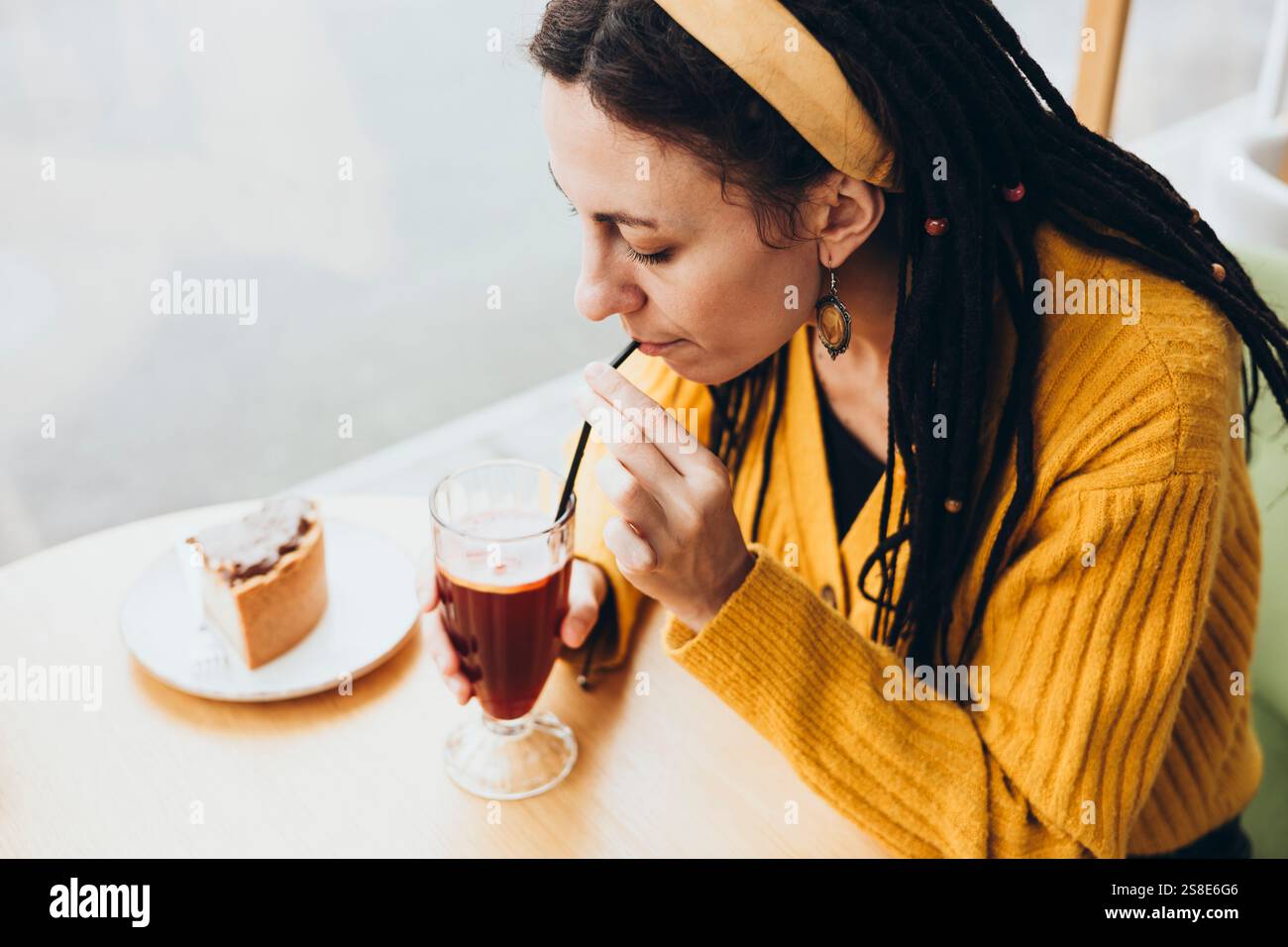 Une femme avec des dreadlocks dans un café sirote une boisson à travers une paille portant un pull jaune et un bandeau, elle aime une tranche de gâteau sur un bois clair Banque D'Images