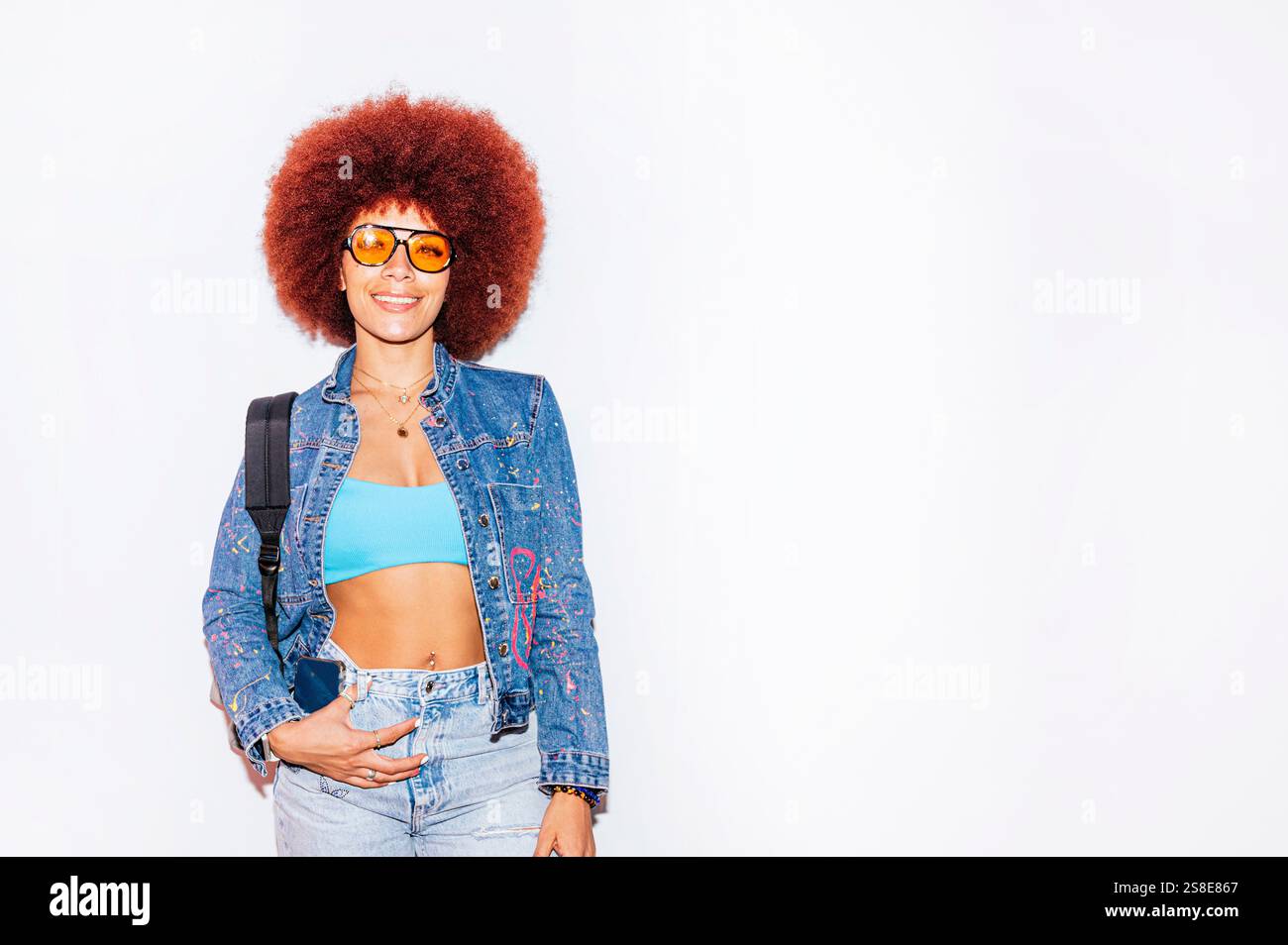 Femme de descendance africaine avec une chevelure afro vibrante, souriante tout en portant des lunettes de soleil orange et une veste en denim, debout contre un backgrou blanc Uni Banque D'Images