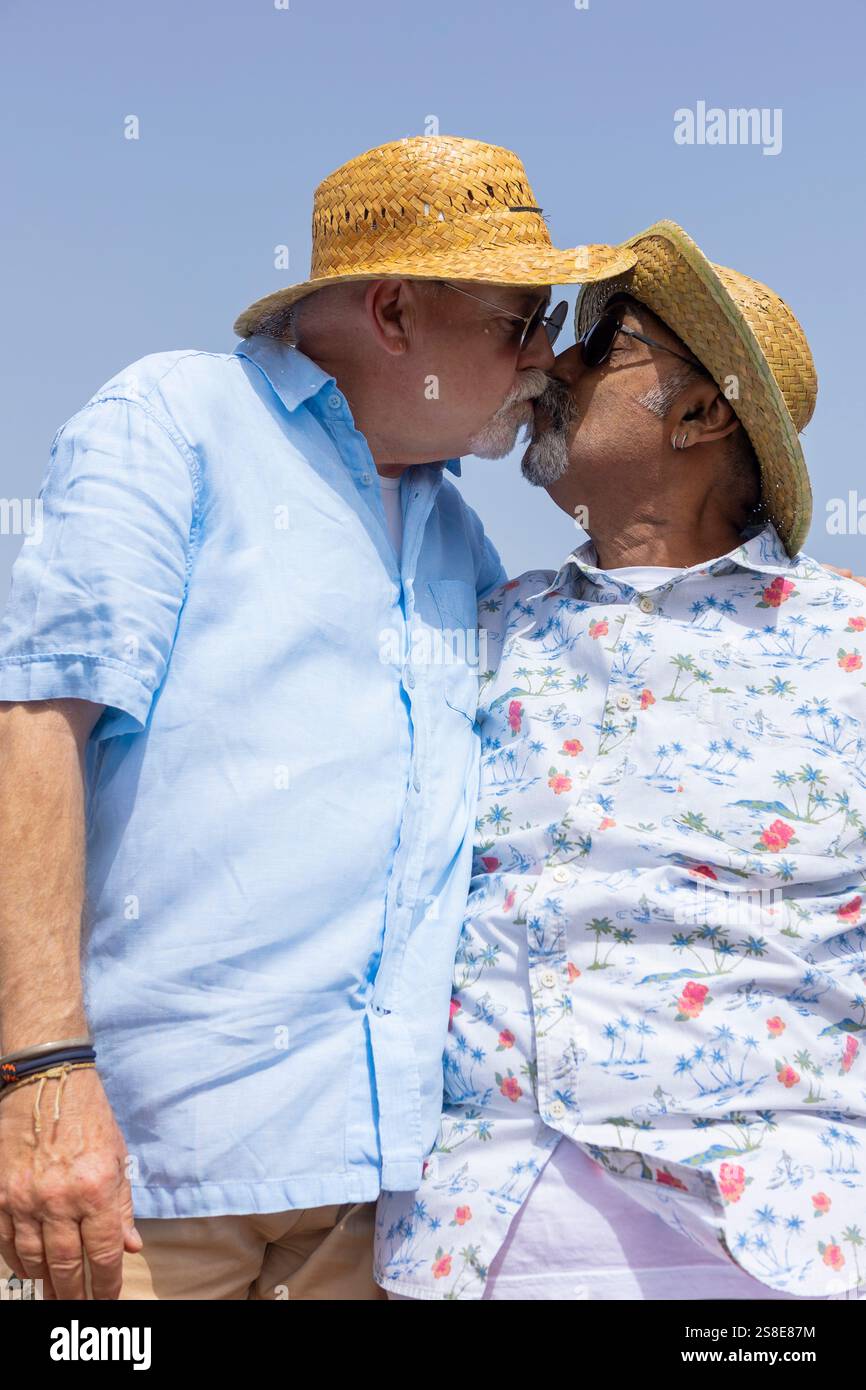 Couple gay senior dans une étreinte tendre, chacun portant des chapeaux de paille et des lunettes de soleil sous un ciel bleu clair. Leur baiser affectueux capture leur amour re Banque D'Images