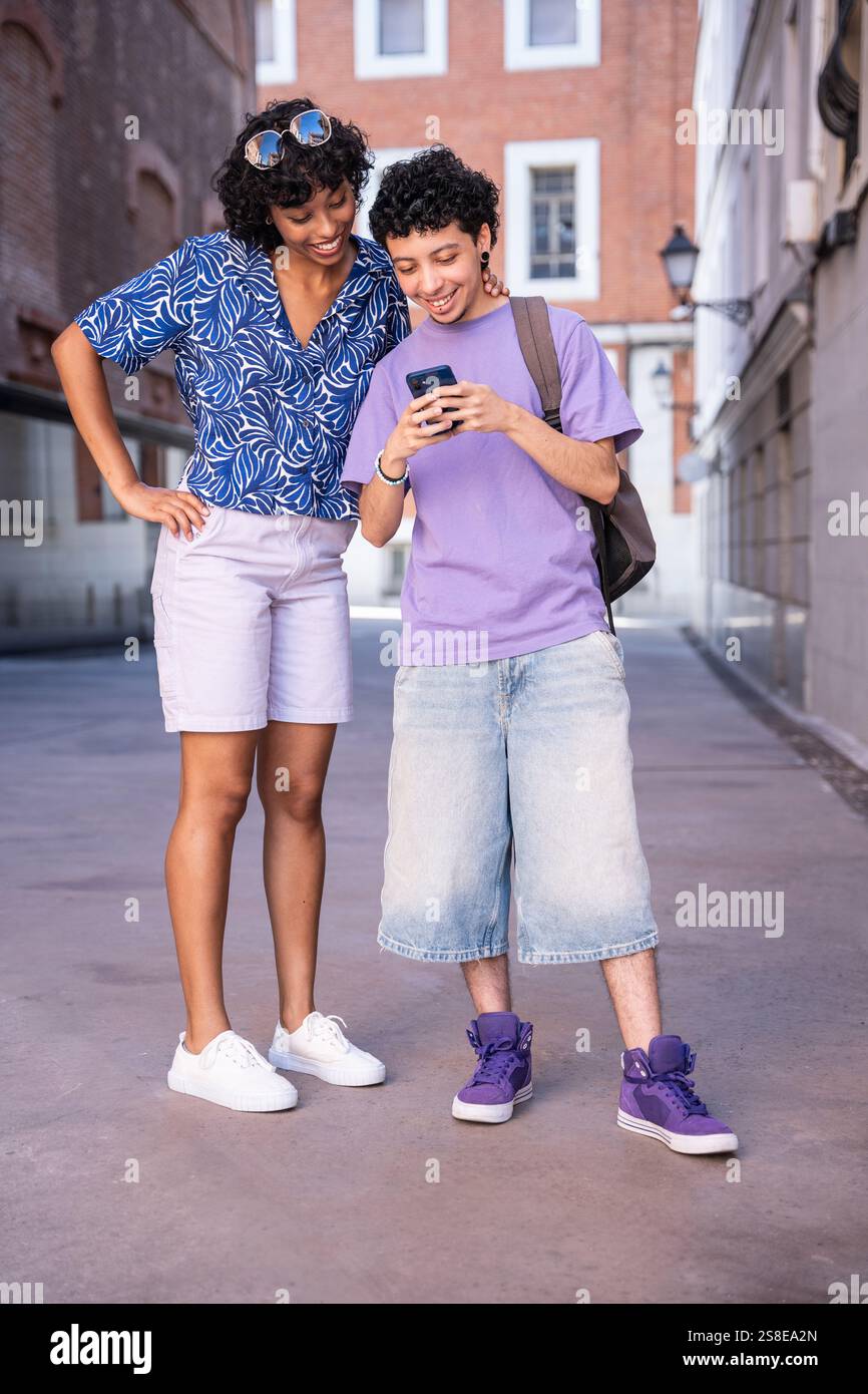 Un couple LGBTQ+ multiethnique partage joyeusement un moment tout en explorant un cadre urbain dynamique. Ils sourient et utilisent un smartphone, entourés de cha Banque D'Images