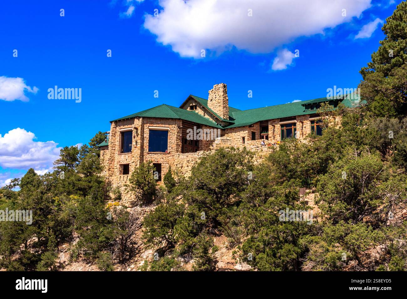 Une grande maison en pierre avec un toit vert se trouve sur une colline. La maison est entourée d'arbres et dispose d'une grande fenêtre sur le devant. Le ciel est clair et bleu, Banque D'Images