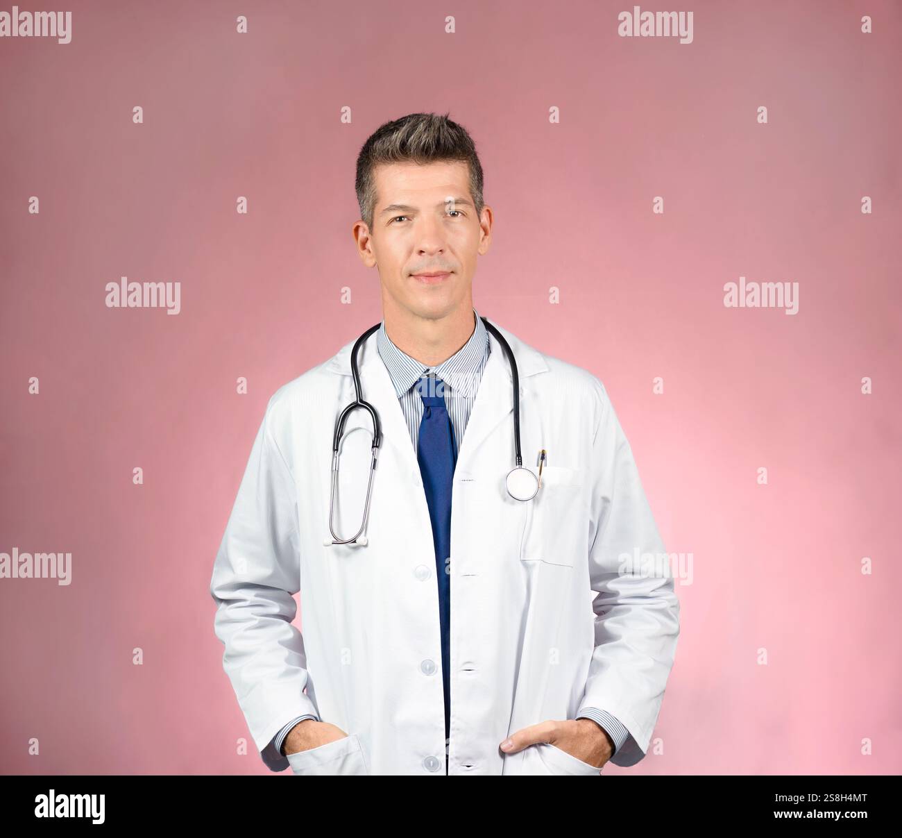 Beau jeune médecin en manteau blanc, stéthoscope autour du cou, cravate et chemise, mains dans les poches de manteau, regardant la caméra. Fond rose Banque D'Images