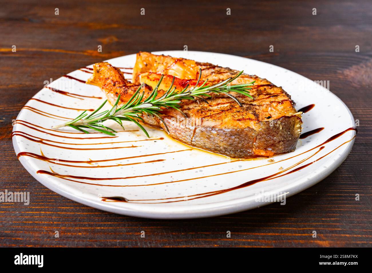 Le steak de saumon grillé repose élégamment sur une assiette blanche, ornée d'un brin de romarin et arrosée de glaçage balsamique. La présentation met en évidence Banque D'Images