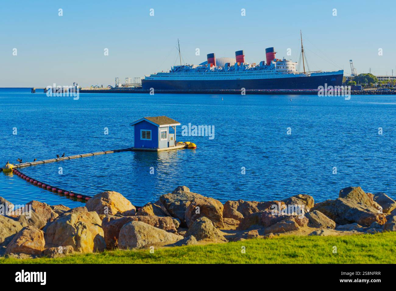 Long Beach, Californie - 15 janvier 2025 : vue sur le navire historique Queen Mary à côté d'une petite maison flottante sur des eaux calmes, entouré de rochers et Banque D'Images