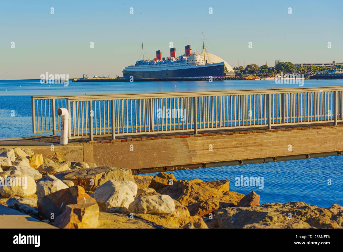 Long Beach, Californie - 15 janvier 2025 : vue depuis une jetée en bois vers le navire Queen Mary, avec une rive rocheuse et des eaux calmes entourant le histo Banque D'Images