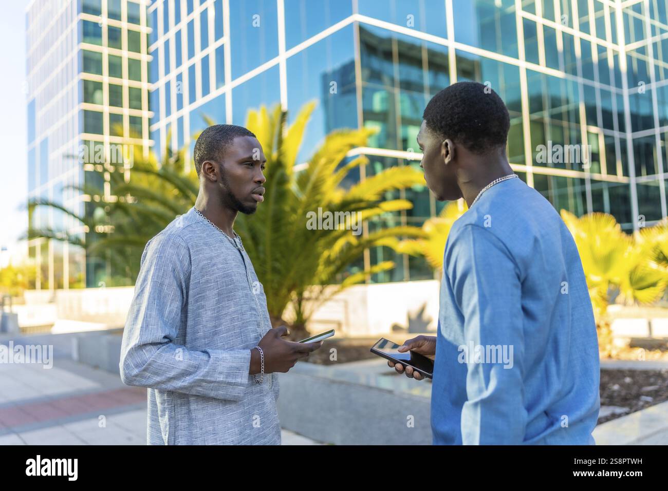 Deux hommes d’affaires sénégalais portant une tenue dashiki traditionnelle utilisent leur smartphone devant un immeuble de bureaux moderne Banque D'Images