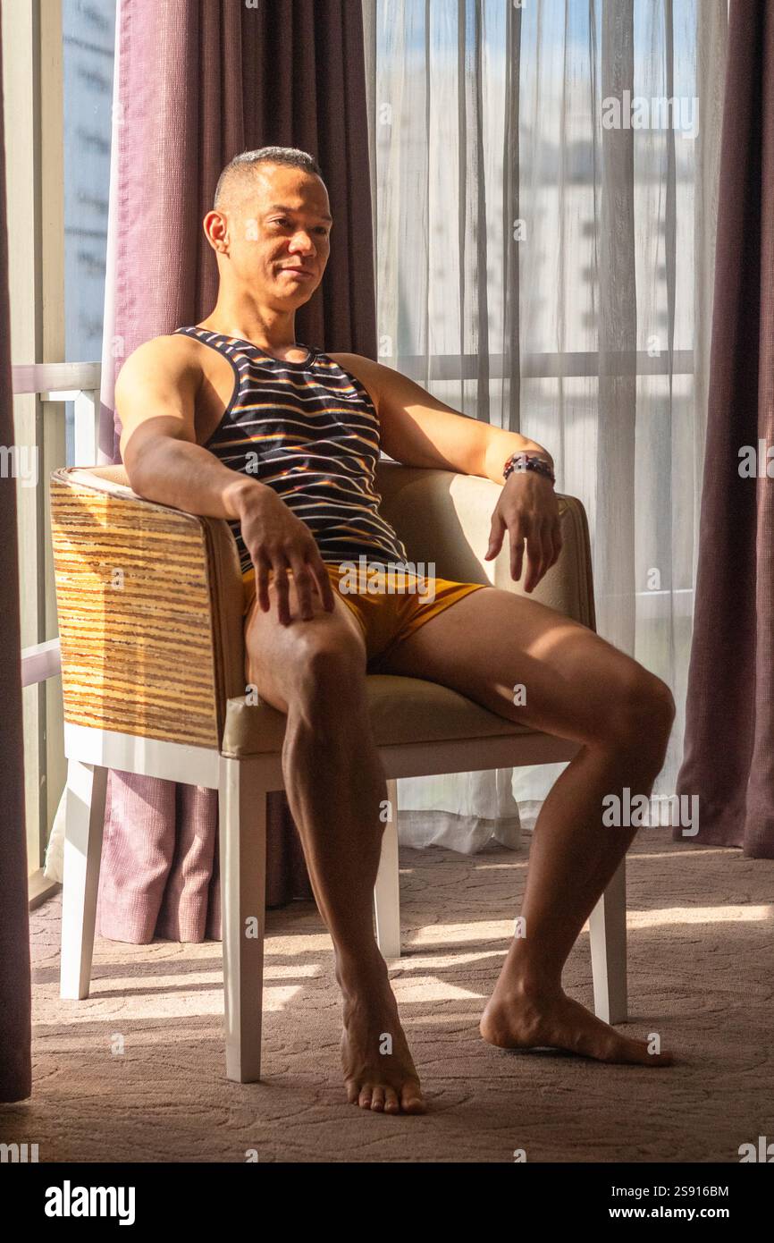 Un beau jeune modèle masculin philippin en short jaune et chemise rayée est assis sur une chaise à Cebu City baigné par la lumière du soleil en fin d'après-midi Banque D'Images