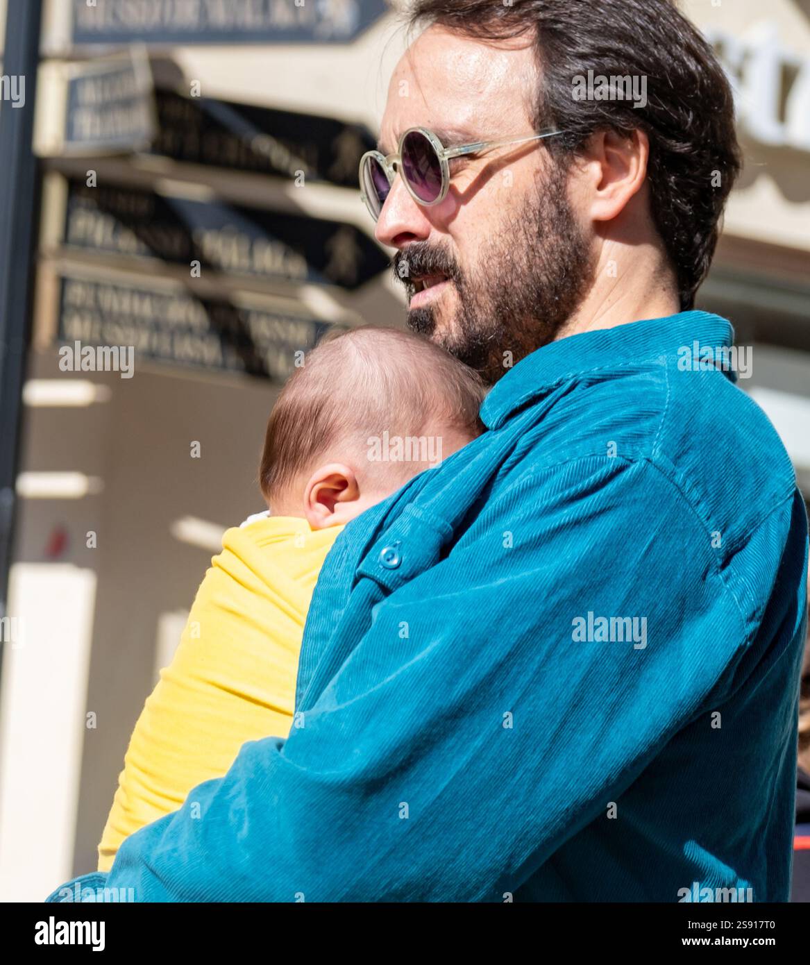 L'image montre un adulte, probablement un homme, portant des lunettes de soleil et une chemise bleue, tenant un bébé. Le bébé semble porter une tenue jaune et Banque D'Images