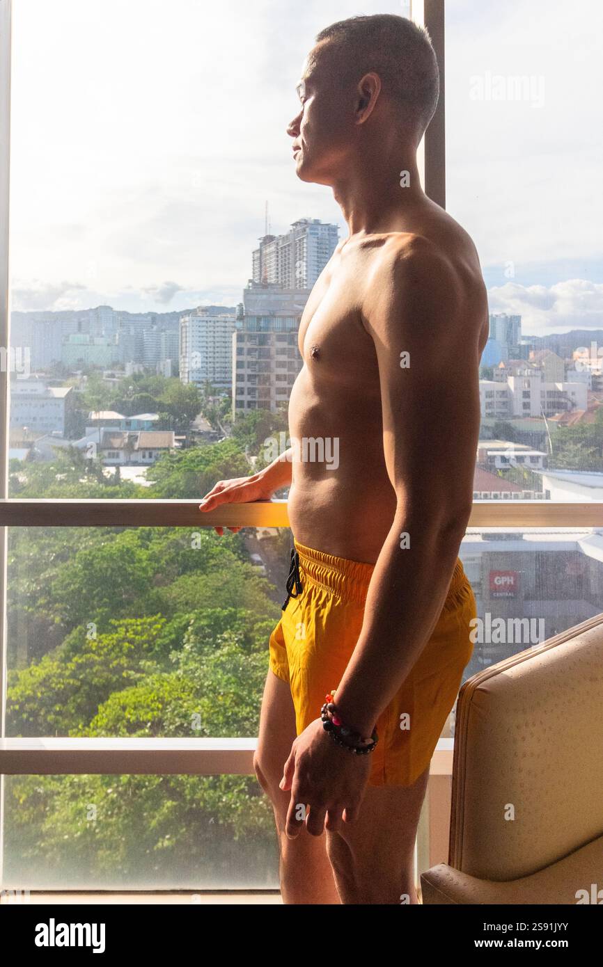 Un beau jeune mannequin masculin philippin en short jaune regarde les gratte-ciel de Cebu City tandis que le soleil chaud de fin d'après-midi coule à travers la fenêtre Banque D'Images Un beau jeune mannequin masculin philippin en short jaune regarde les gratte-ciel de Cebu City tandis que le soleil chaud de fin d'après-midi coule à travers la fenêtre Banque D'Images