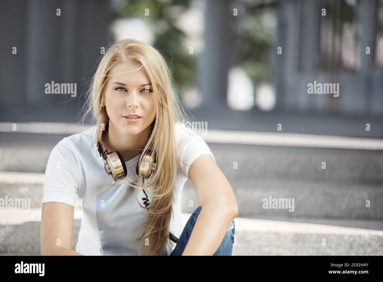 Belle jeune femme avec des écouteurs de musique vintage autour de son cou, assise devant un fond urbain et regardant droit devant Banque D'Images