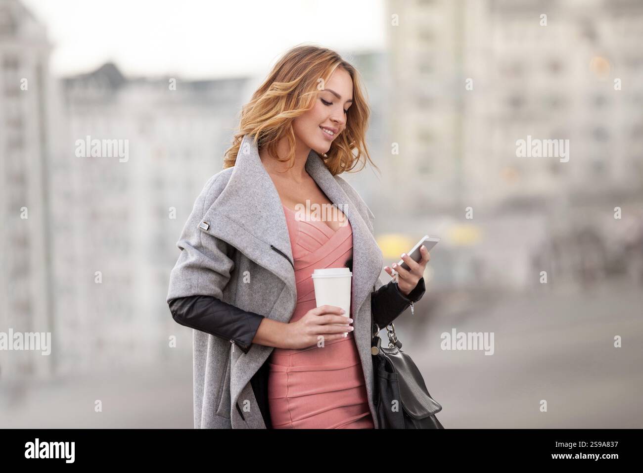 Une femme d'affaires vérifiant ses e-mails sur son téléphone portable et tenant une tasse de café dans sa main devant un fond urbain Banque D'Images