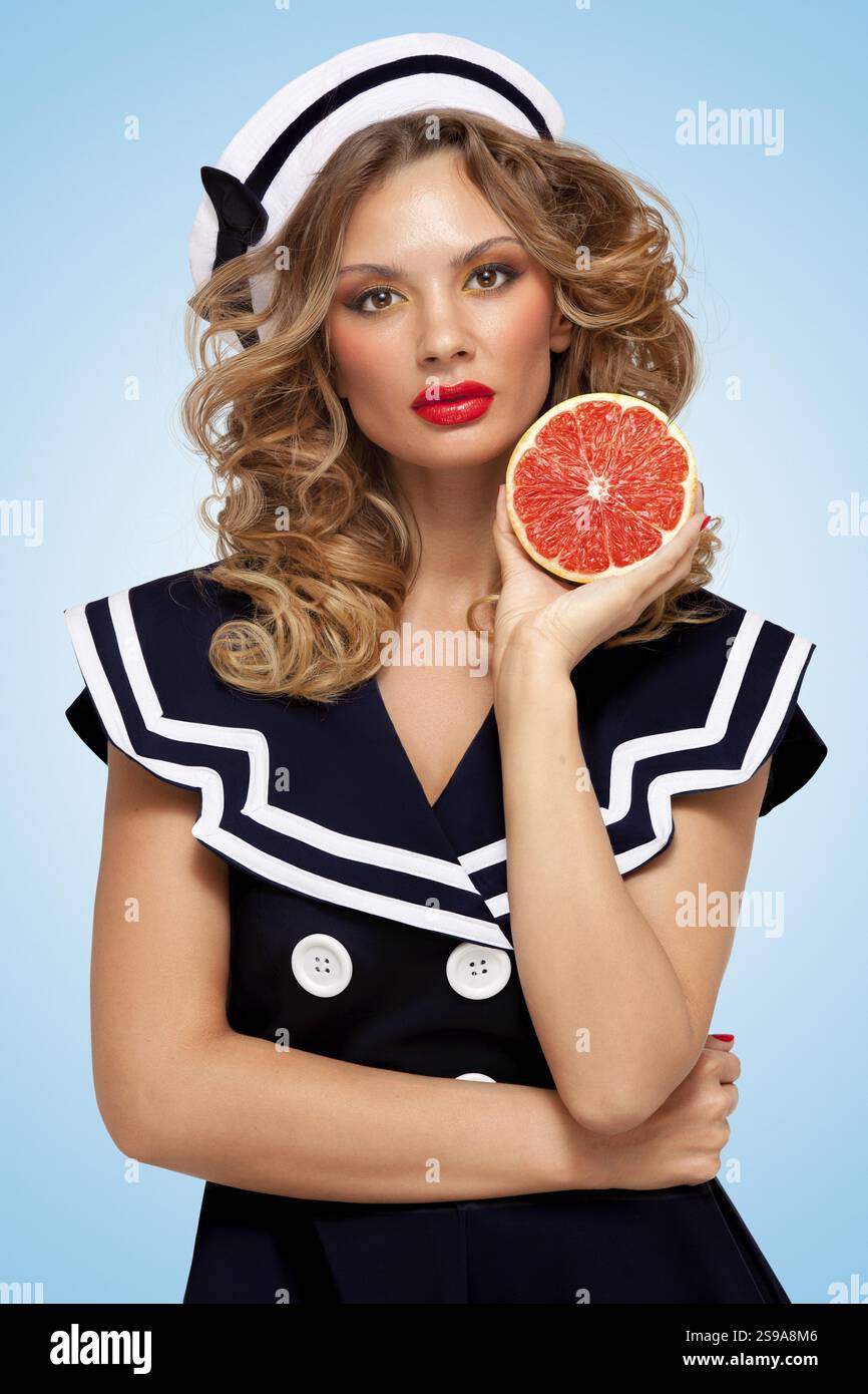 Belle jeune femme dans une robe de marin tenant un pamplemousse rouge, soin de la peau bio anti vieillissement pour le corps et le visage sur un fond bleu Banque D'Images