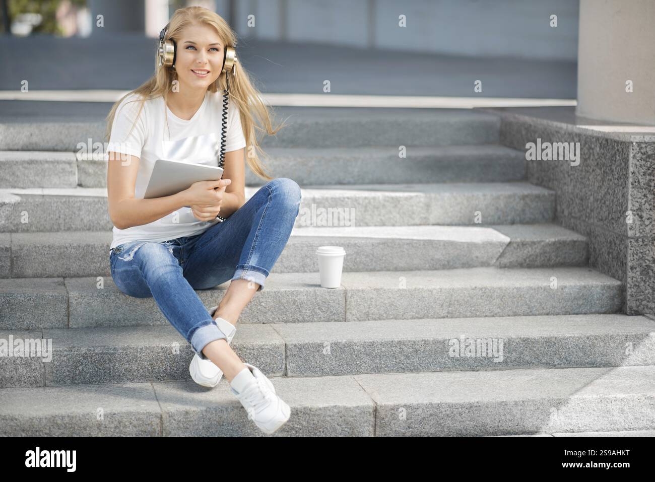 Jeune femme heureuse avec des écouteurs vintage et une tasse de café à emporter, surfant sur Internet sur une tablette PC, écoutant de la musique et assis sur un escalier Banque D'Images