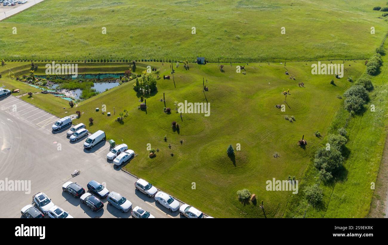 Vue aérienne d'un parc herbeux avec des sculptures et un petit étang, adjacent à un parking avec plusieurs voitures. Le paysage est entouré de f ouvert Banque D'Images