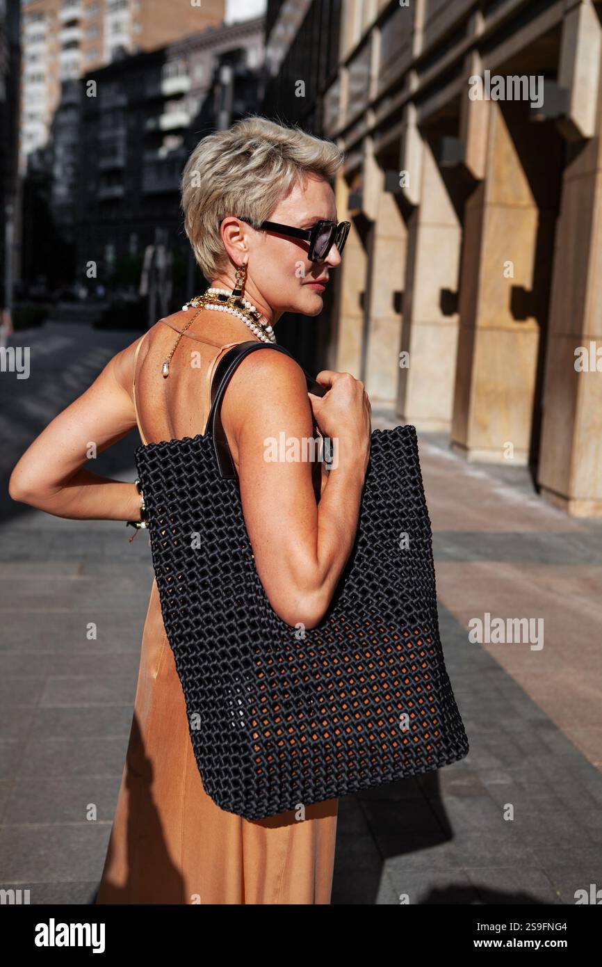 Femme blonde à la mode avec les cheveux courts dans une tenue élégante avec sac tissé noir, robe de soleil beige d'été avec dos ouvert et bijoux uniques, Aga debout Banque D'Images