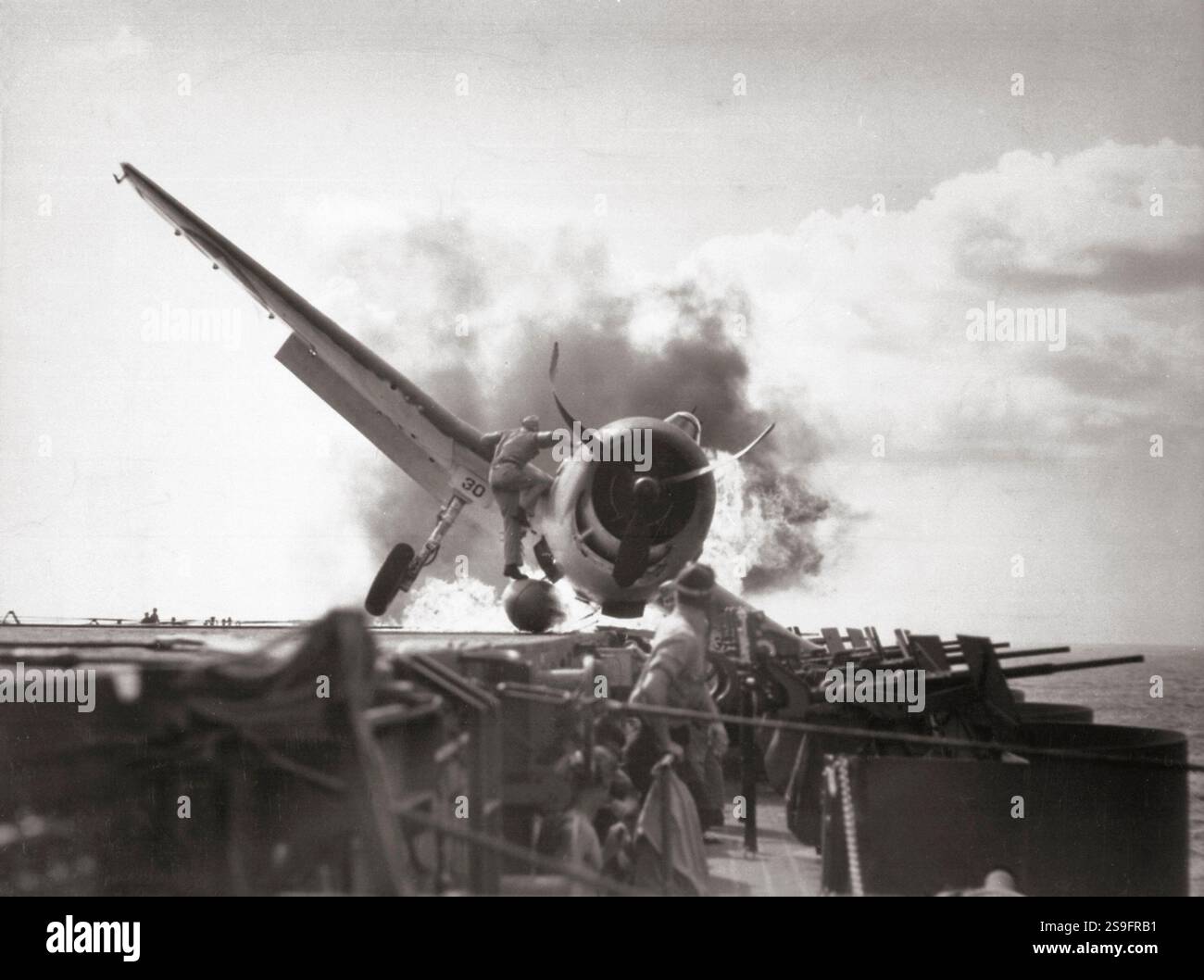 Atterrissage écrasé d'un Grumman F6F-3 Hellcat (numéro 30) du Fighting Squadron 2 (VF-2) à bord du porte-avions USS Enterprise (CV-6), dans la galerie de canons de 20 mm côté bâbord du porte-avions, le 10 novembre 1943. Le lieutenant Walter L. Chewning, Jr., USNR, l'officier de catapulte, est vu grimper sur le côté de l'avion pour aider le pilote à échapper à l'avion en feu. Le pilote, l'enseigne Byron M. Johnson, s'est échappé sans blessure grave. L'Enterprise était alors en route pour soutenir l'opération Gilberts. Notez le réservoir de carburant ventral rompu de l'avion. Banque D'Images