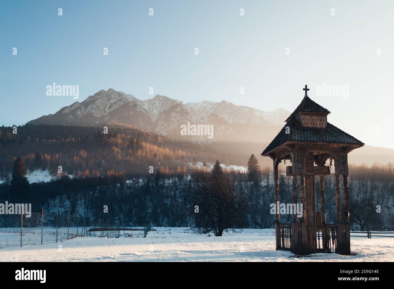 Une scène hivernale sereine mettant en vedette un pavillon roumain traditionnel en bois avec une croix, contre les montagnes enneigées de Piatra Craiului. Le golde Banque D'Images