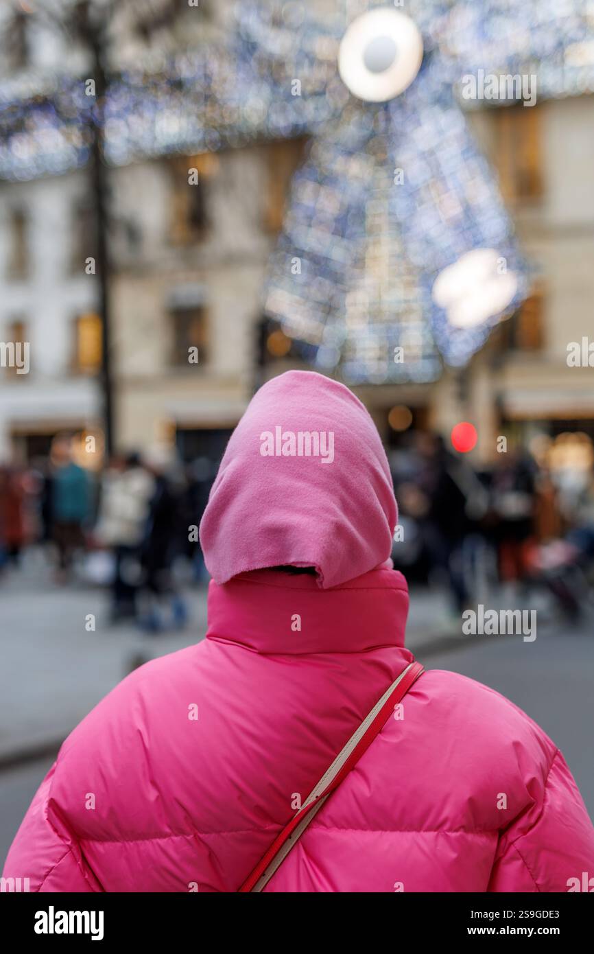 Une personne en veste à capuche rose se tient devant le magasin phare Chanel à Paris pendant la saison de Noël 2024, avec des lumières et un décor festifs Banque D'Images