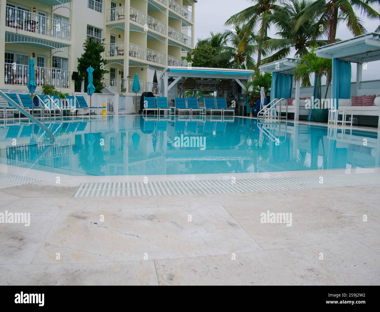Basse vue large sur une terrasse de la piscine vers une piscine d'eau bleue calme piscine avec cabanas, chaises longues, parasols, palmiers verts et bâtiments dans le bac Banque D'Images Basse vue large sur une terrasse de la piscine vers une piscine d'eau bleue calme piscine avec cabanas, chaises longues, parasols, palmiers verts et bâtiments dans le bac Banque D'Images
