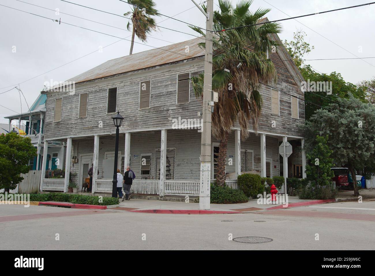 Vieille maison de deux étages en bois vieilli avec une grande enveloppe autour du porche avant. Toiture en métal rouillé. Bouche d'incendie sur la droite. Palmiers et buissons verts Banque D'Images