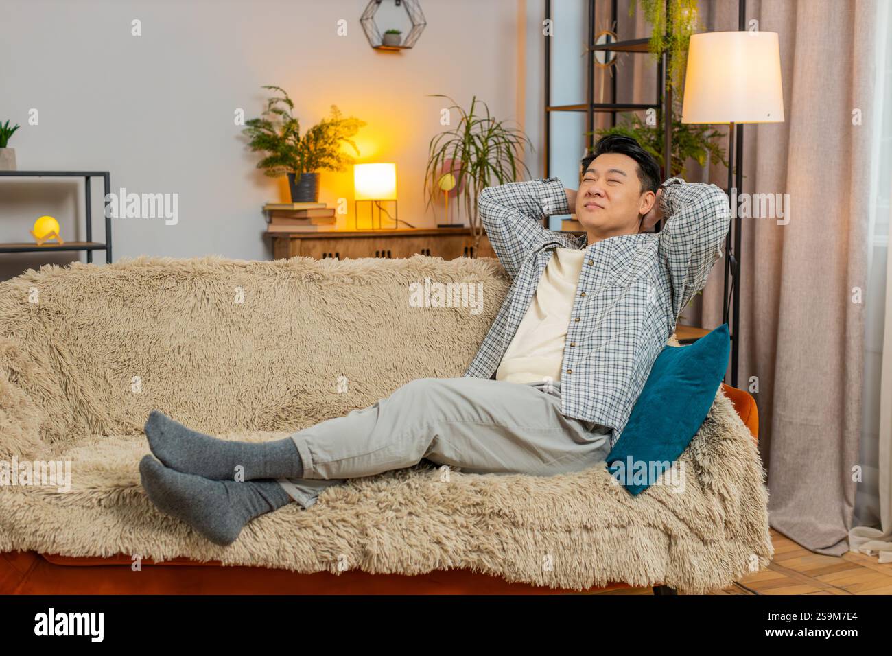 Heureux homme chinois d'âge moyen insouciant assis confortablement avec la main derrière la tête sur le canapé dans le salon à la maison. Souriant mec coréen se relaxant sur le canapé pensant regarder autour dans la chambre de l'appartement. Banque D'Images