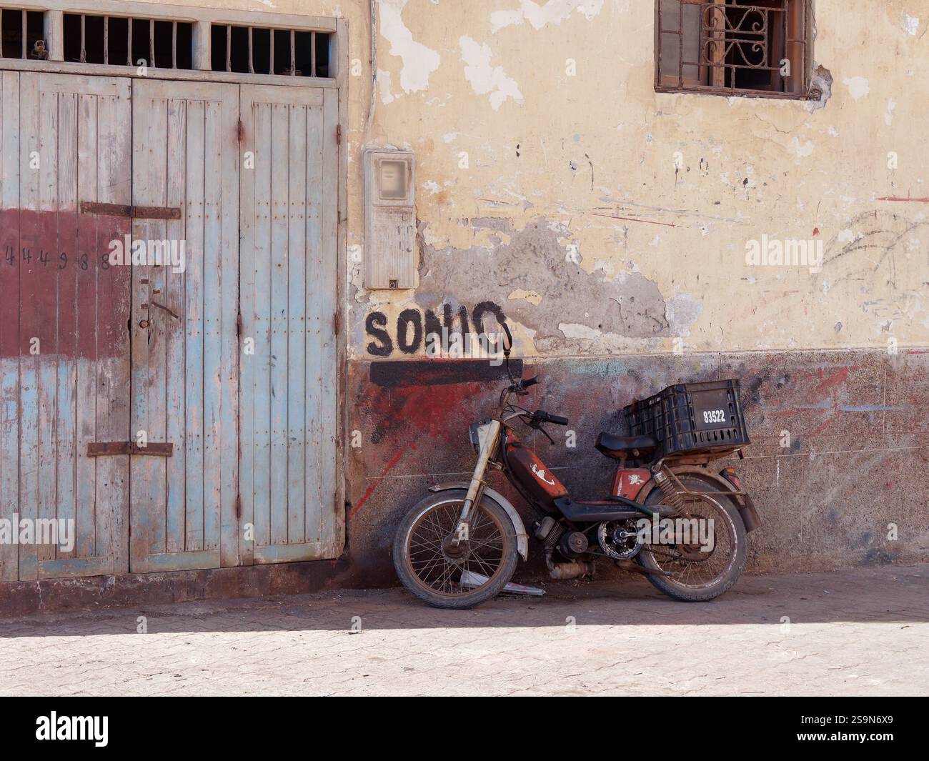Vieille moto appuyée contre un bâtiment délabré à porte bleue à Essaouira, Maroc. 26 janvier 2025 Banque D'Images