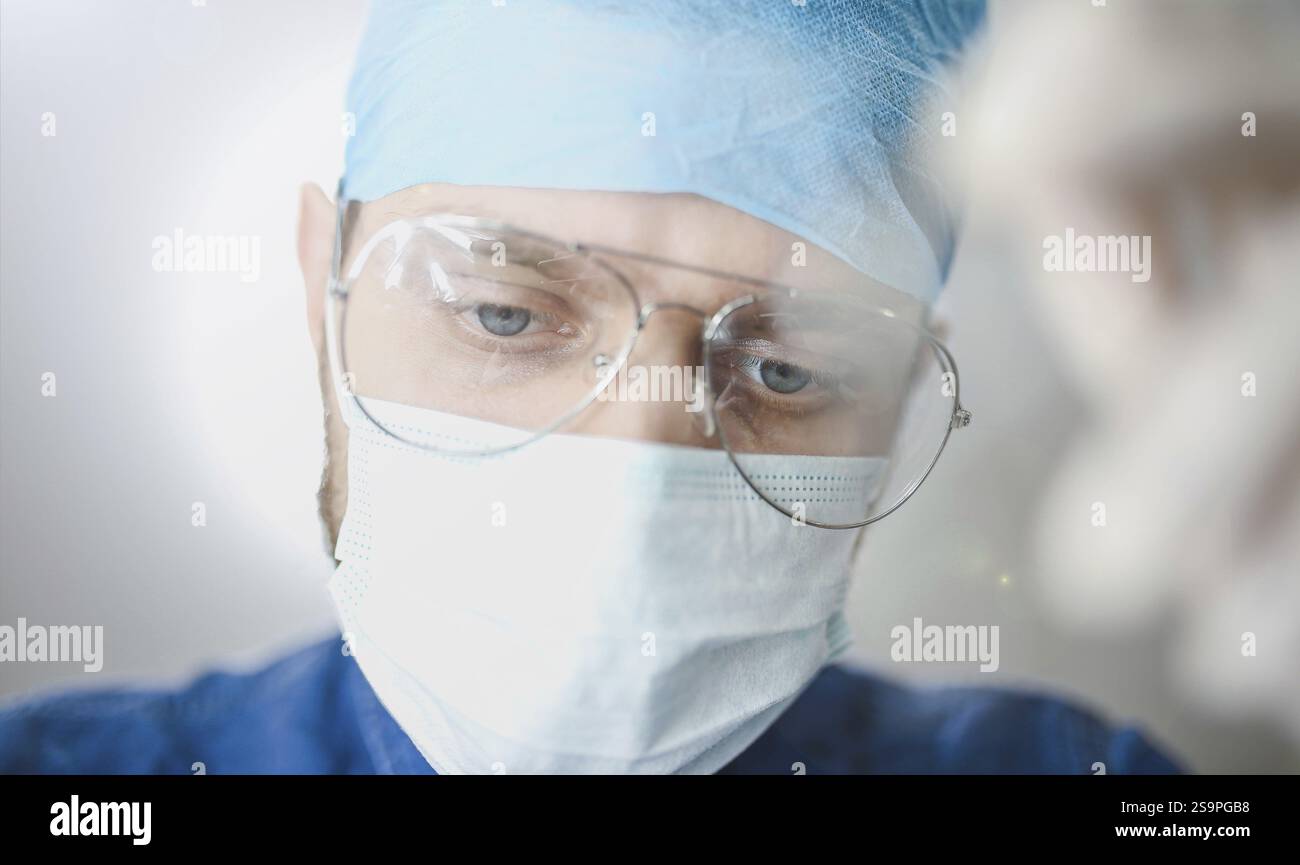 Gros plan d'un médecin masculin portant des lunettes, un masque chirurgical et des gommages bleus, axé sur une tâche Banque D'Images