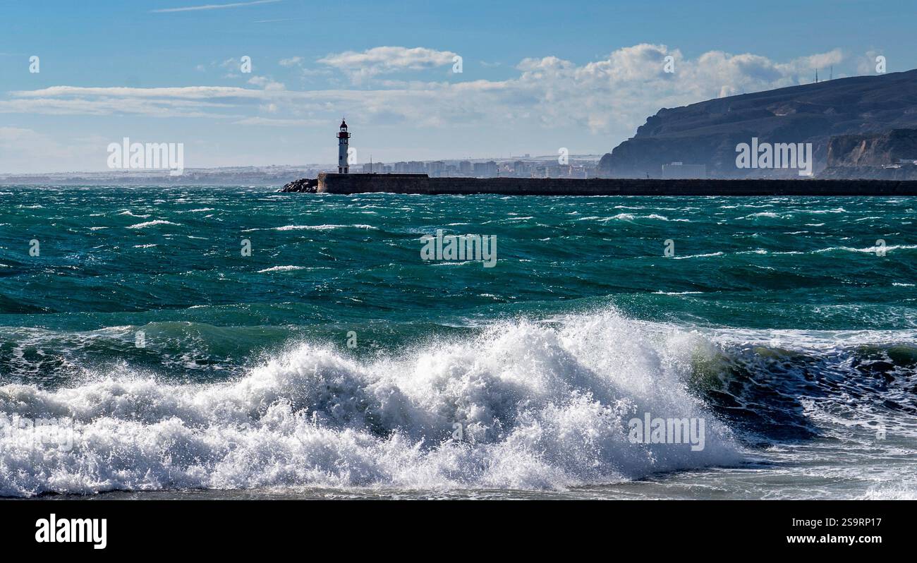Une vue panoramique sur un phare au bord de la mer, avec des vagues s'écrasant contre le rivage. Le ciel est partiellement nuageux, et un paysage côtier est visible dans le b. Banque D'Images