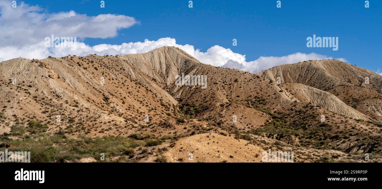 Une vue panoramique sur les montagnes escarpées avec des pentes sablonneuses sèches sous un ciel bleu vif. Le paysage présente une végétation clairsemée et une forma nuageuse spectaculaire Banque D'Images