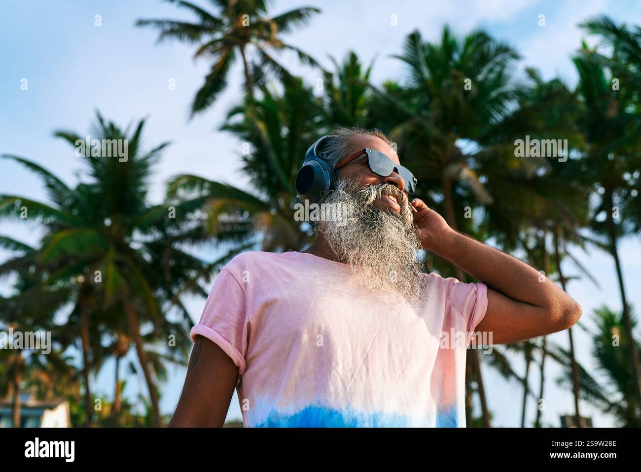 L'homme plus âgé avec la barbe porte des écouteurs, des lunettes de soleil près des palmiers. Profitez de la musique. Essaie peut-être la suppression active du bruit. Cadre tropical Banque D'Images