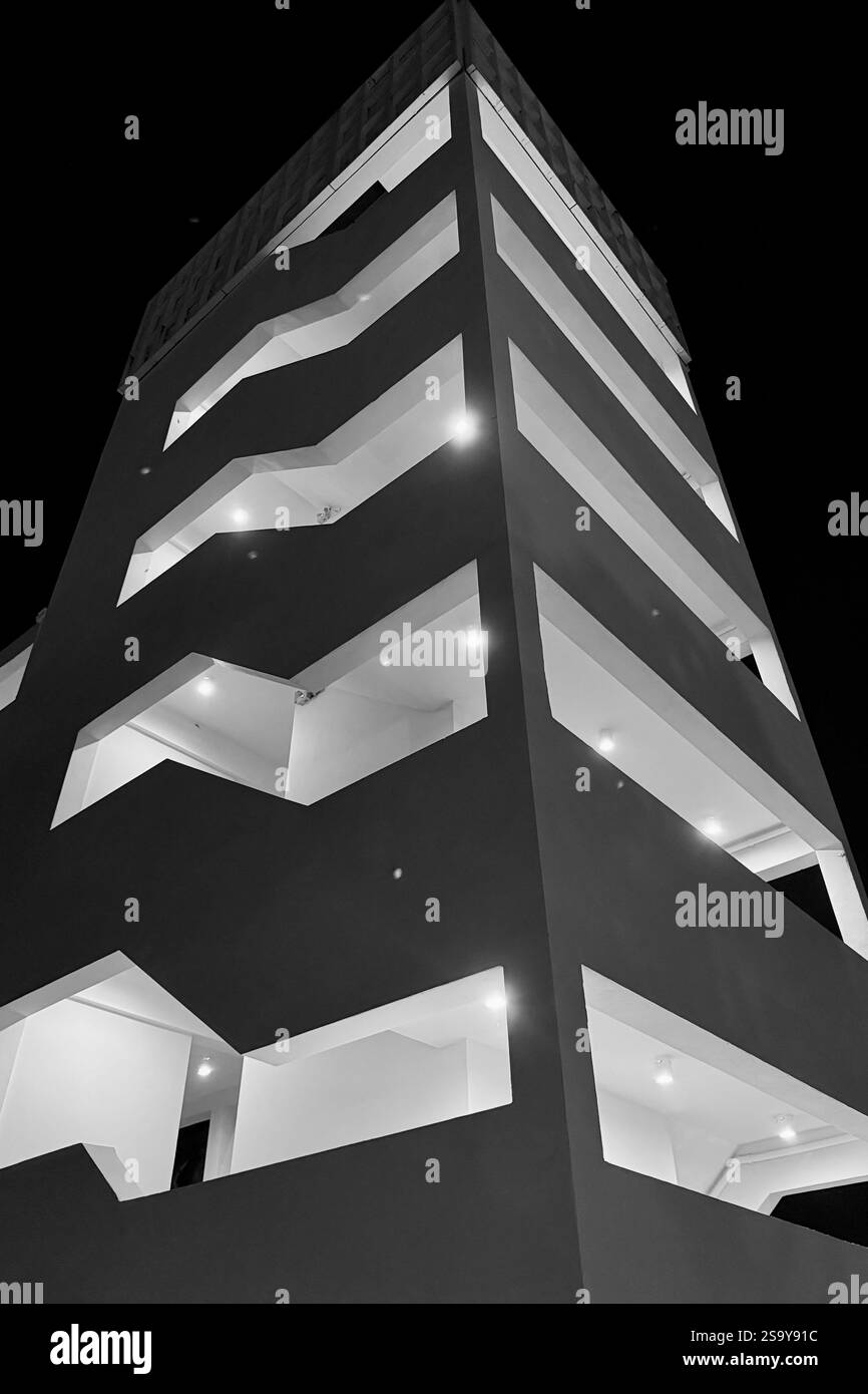 Bâtiment moderne la nuit Une vue en angle bas d'un bâtiment moderne avec des motifs géométriques et des lumières vives, capturée la nuit en noir et blanc. - Image de stock capturée avec un smartphone