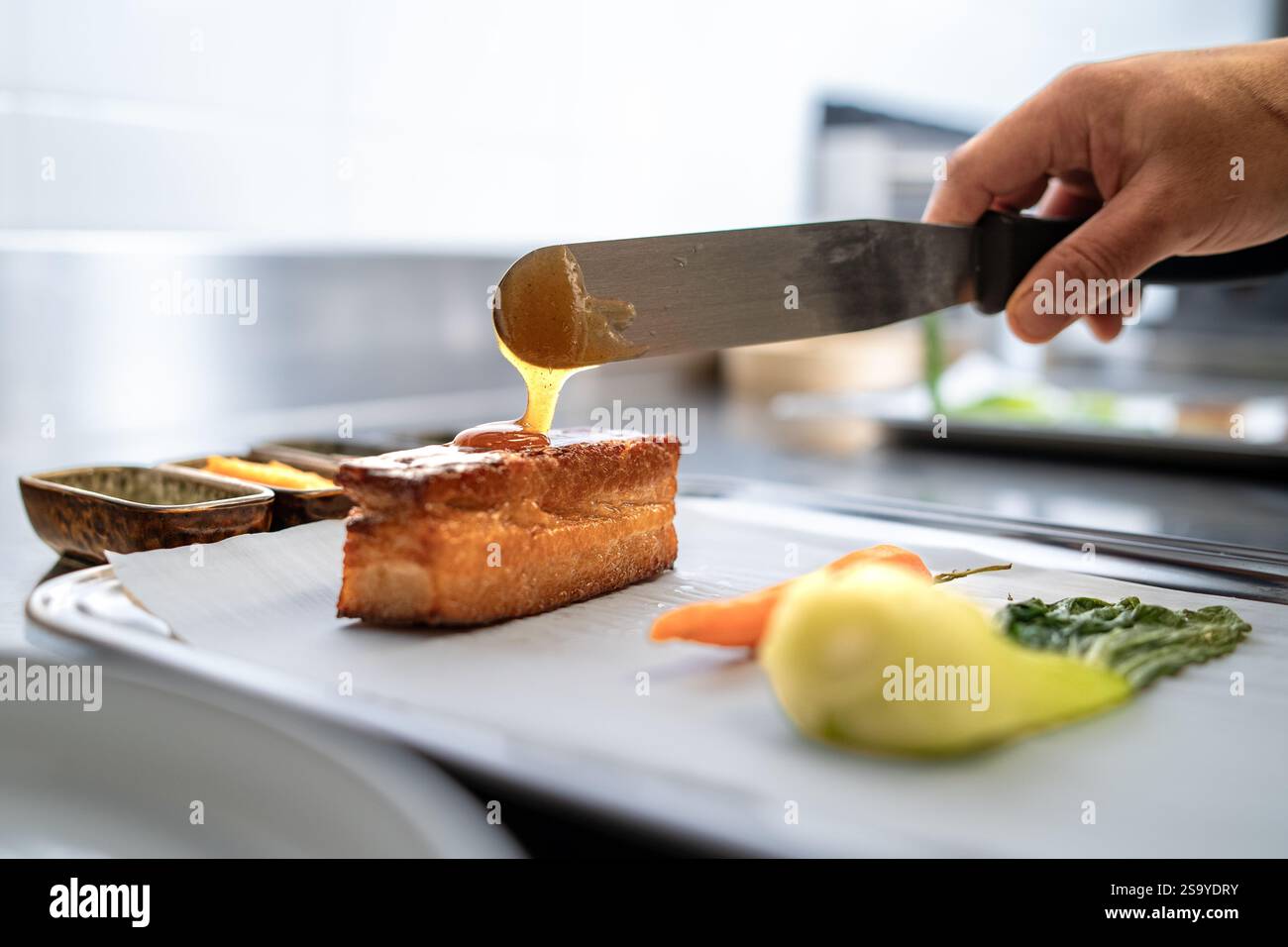 Chef versant un glaçage doré brillant sur un ventre de porc croustillant parfaitement cuit, accompagné de légumes frais, dans un cadre de cuisine professionnel. Banque D'Images