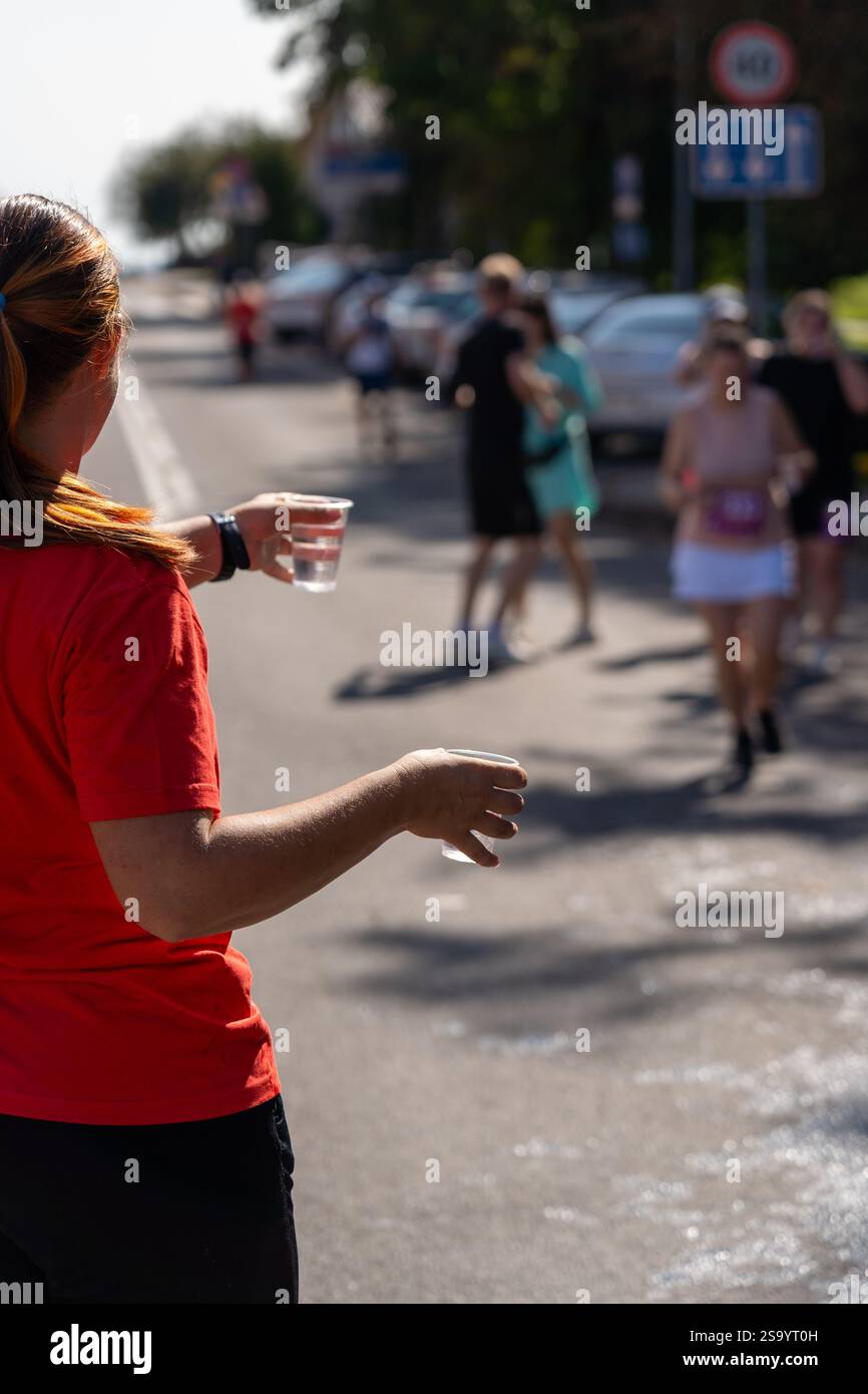 Un volontaire portant un t-shirt rouge tient une tasse d'eau en plastique à un coureur de marathon. L'image capture l'esprit de soutien et de communauté à un sport Banque D'Images