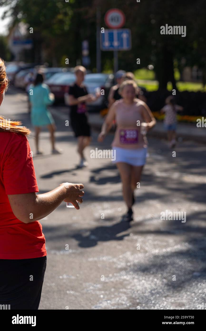Un volontaire portant un t-shirt rouge tient une tasse d'eau en plastique à un coureur de marathon. L'image capture l'esprit de soutien et de communauté à un sport Banque D'Images