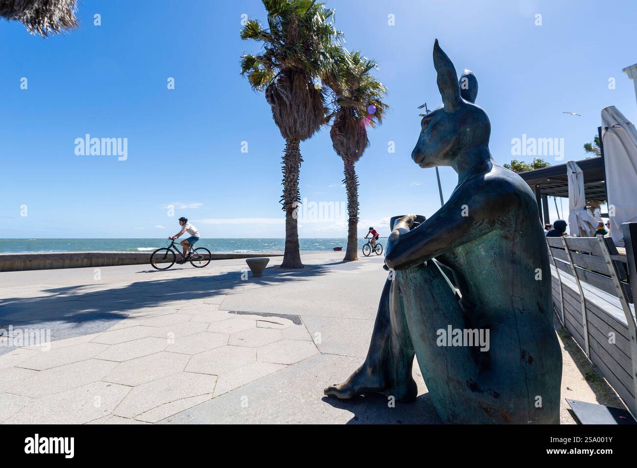 Une statue de lapin en bronze avec une tasse de café surveille la mer sur une promenade ensoleillée de St Kilda tandis que les cyclistes passent devant des palmiers sous le ciel bleu. Banque D'Images