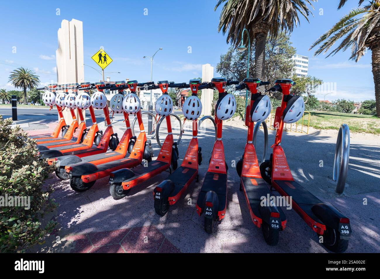 Un groupe de scooters électriques orange alignés dans une rangée avec des casques blancs à louer sur le soleil, ciel bleu jour d'été, St Kilda promenade, Melbourne. Banque D'Images
