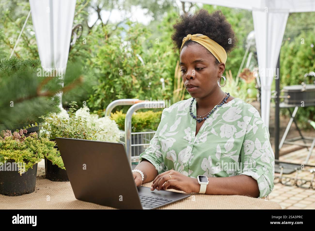 Prise de vue sur la poitrine d'une paysagiste afro-américaine concentrée tapant sur un ordinateur portable, tout en travaillant sur un projet de jardin en ligne assis à table avec une verdure estivale animée à l'extérieur Banque D'Images