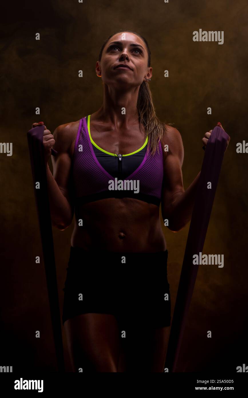 Une jeune femme concentrée présente un entraînement de force physique en utilisant des bandes de résistance dans un cadre de studio intense avec un fond sombre et vibrant. Banque D'Images
