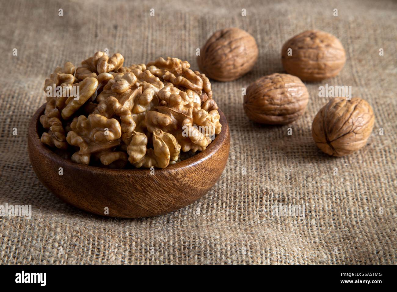 Un bol en bois rempli de cerneaux de noix, placé à côté des noix entières dans leurs coquilles sur toile de jute Banque D'Images