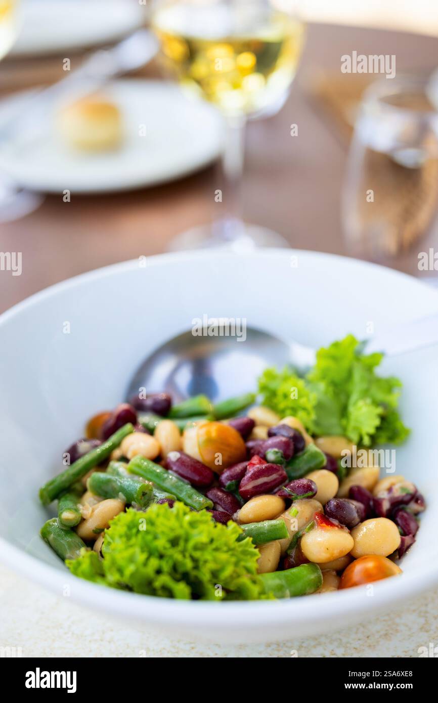 Une salade de haricots frais mélangée servie dans un bol blanc, garnie de laitue verte et de tomates cerises. Un verre de vin blanc et un cadre de table complètent le restaurant Banque D'Images