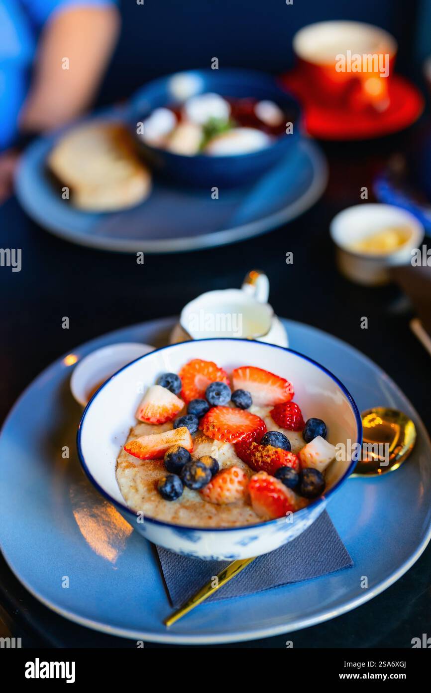 Flocons d'avoine chauds servis dans un bol blanc et bleu, garnis de fraises fraîches, de myrtilles et de crème sur assiette bleue. Banque D'Images