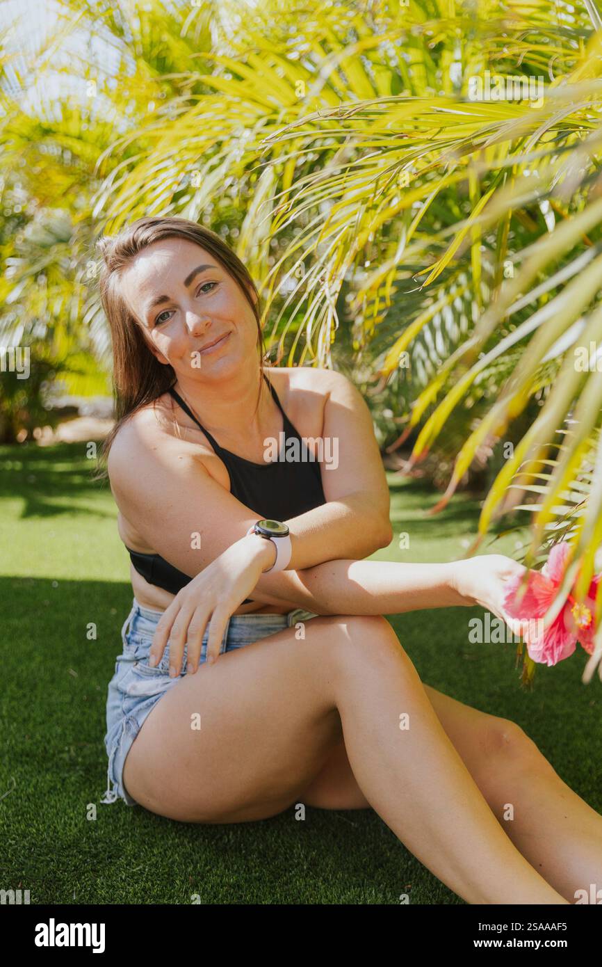 Une femme est assise sur l'herbe avec une fleur rose à la main. Elle porte un débardeur noir et un short bleu Banque D'Images