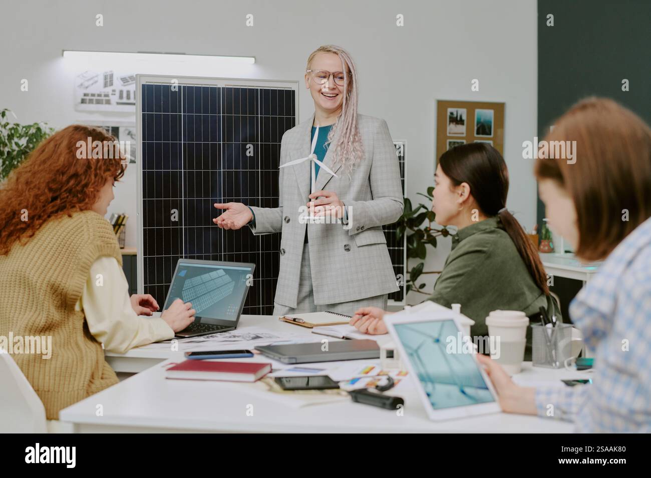 Femme d'affaires souriante donnant une présentation dans un cadre de bureau moderne avec des collègues engagés et discutant à table. La présentation comprend l'affichage de panneau solaire et divers appareils technologiques Banque D'Images