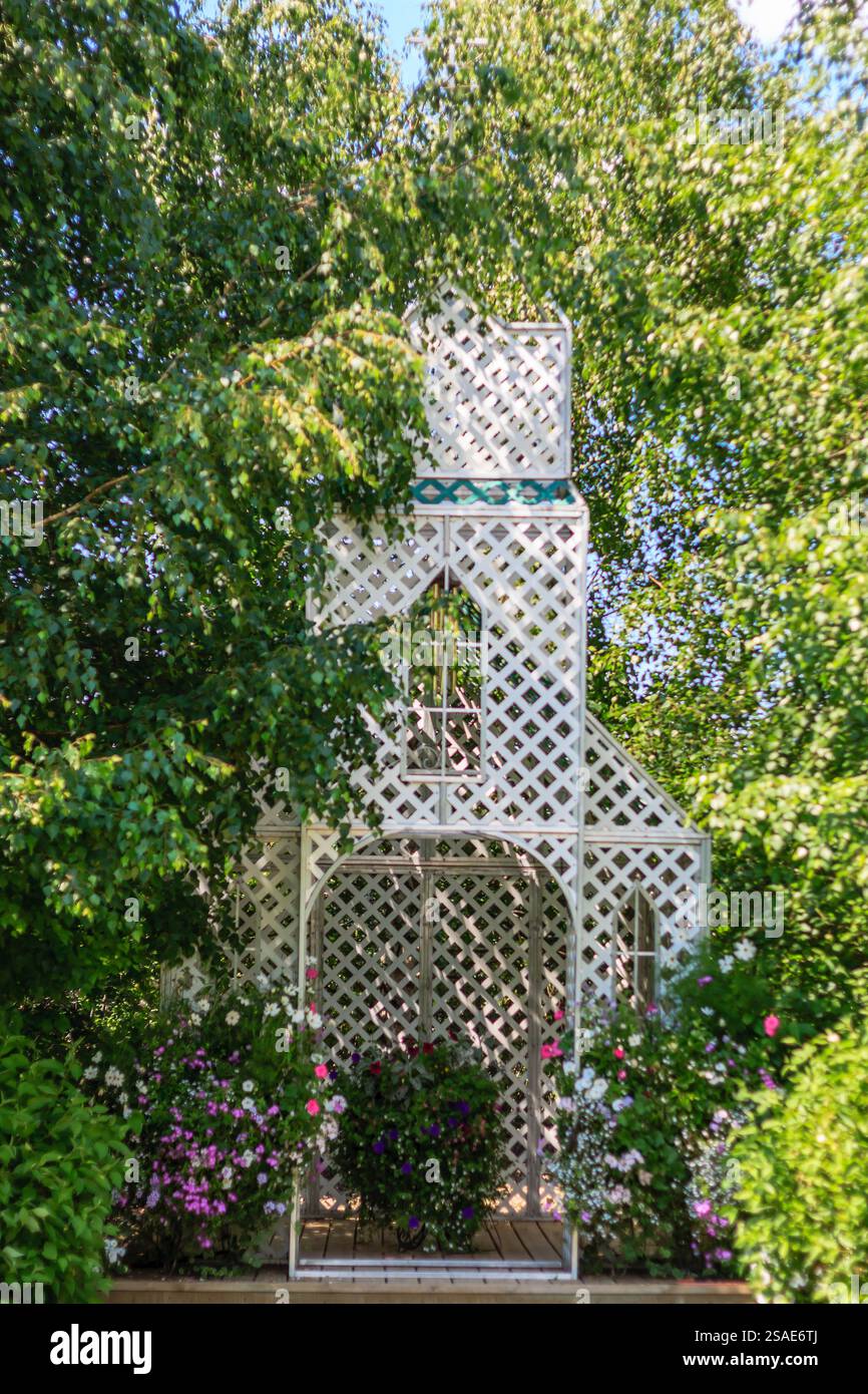 Un gazebo blanc avec un toit en treillis se trouve dans un jardin verdoyant luxuriant. Le belvédère est entouré d'une variété de fleurs et de plantes, créant un pois serein Banque D'Images