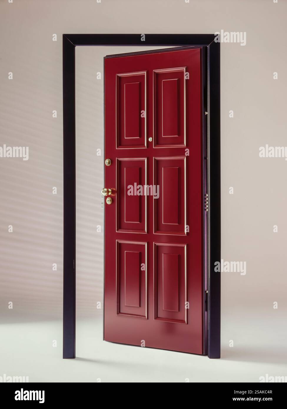 Porte blindée de sécurité. Une porte rouge classique légèrement entrouverte dans un cadre noir, projetant de douces ombres diagonales sur un mur blanc cassé dans un éclairage minimaliste Banque D'Images